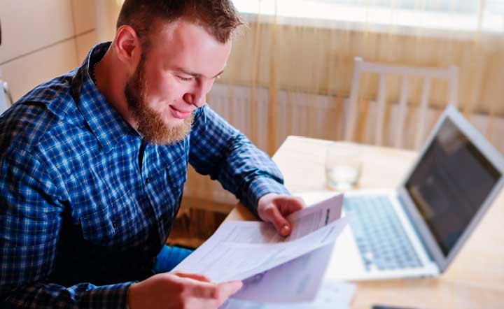Bearded man sitting at a kitchen table, looking down at tax forms, a laptop open in front of him