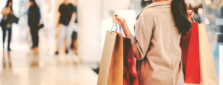 A woman wearing a tan coat, carrying shopping bags walks through a mall.