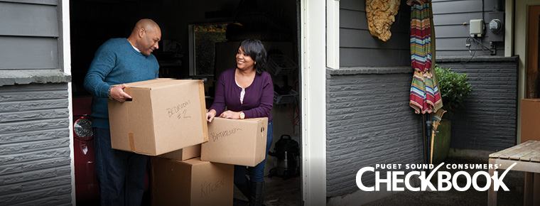 A man and woman stand in their garage doorway. The man is carrying a cardboard box labeled "BEDROOM # 2" in black marker. A woman, who is smiling at him, rests her hands on top of a stack of labeled cardboard boxes. A white text logo in the lower right says "Puget Sound Consumers' Checkbook."