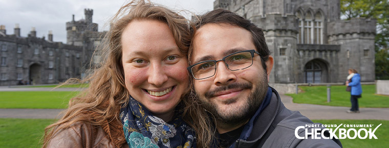 Couple huddled close together, smiling for the camera while situated in front of a medieval castle. Consumers' Checkbook logo in bottom right corner.