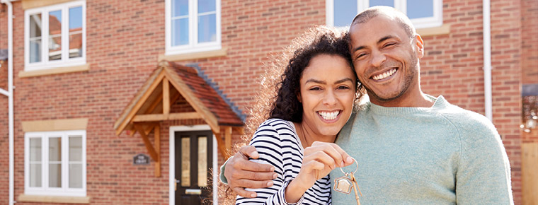 Image of a young, smiling couple standing in front of a brick home with one of the individuals holding house keys in their hand.