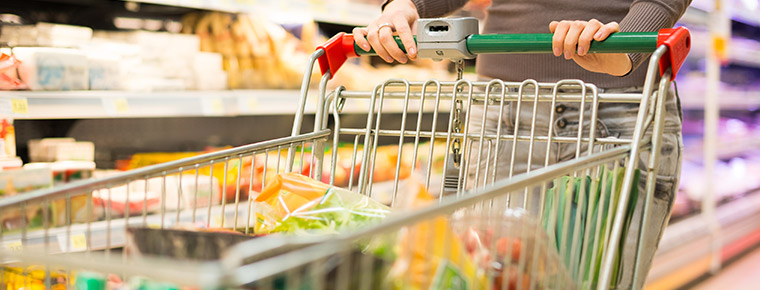 A person pushes a grocery cart down the dairy aisle of the grocery store. The cart contains lots of fresh vegetables and other items. The person's hands and lower body are visible.
