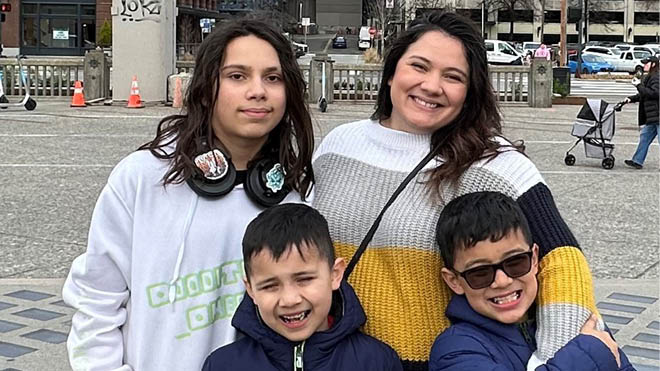 This is a photo of Marybeth, BECU employee, and her three sons. They are all smiling for a group photo. Marybeth has her arms wrapped around them and is wearing a stripped sweater. They are outside and behind them is a parking lot with cars parked. 