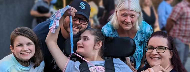 Young girl in a wheelchair smiles as she looks back at four other members of her family, who are surrounding the young girl on all sides while posing and smiling for the photo.