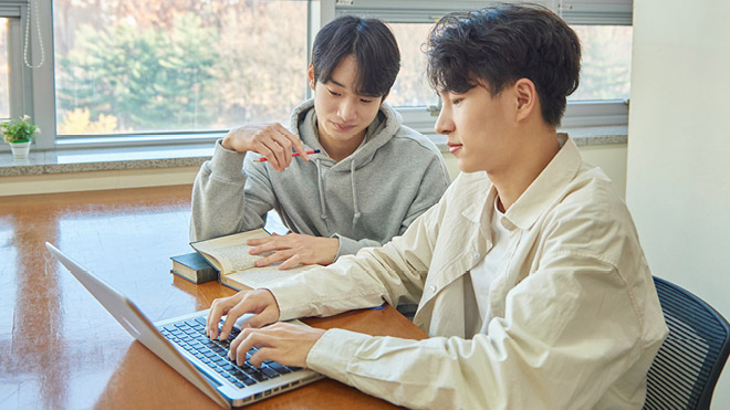 Two young college-aged students sitting at a table inside located in front of large windows. Trees are in the background outside. The two students are looking together at the screen of a laptop that's in front of one of the students, who is typing on the laptop keyboard. The other student not in front of the laptop is holding a pen in one hand and has a book in their other hand resting on the table, open to a page.