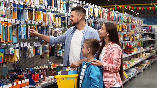 A young boy stands next to his mother and father in the aisle of an office supplies store. The family is facing a wall full of writing utensils and other back-to-school supplies, with the father reaching to pull an item off the shelf.