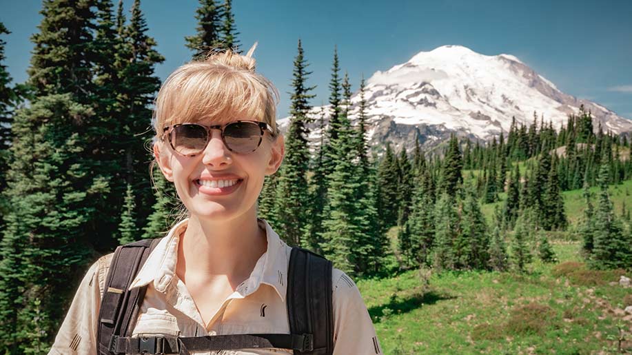 A woman wearing sunglasses, a collared shirt and a backpack smiles at the camera with evergreen trees, Mount Rainier and blue sky in the background.
