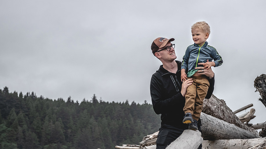 A man holds a child standing up on driftwood at a Washington beach. They're wearing sweatshirts and the sky is cloudy. Ocean mist drifts across the evergreen trees in the background.