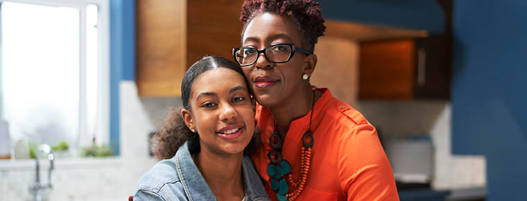 A mother and her teen daughter stand together in their kitchen facing the camera for a photo. The mom has her arm around the teen and is resting her cheek against her daughter's head.