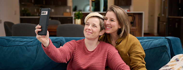 Two women sit close together on couch, holding hands. Woman on the left is holding phone up to take a selfie.