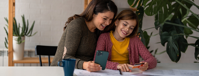 Mother holding a mobile phone sits next to her child who is holding a credit card. They are sitting at a table, smiling. A large green plant is in the background.
