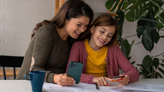 Mother holding a mobile phone sits next to her child who is holding a credit card. They are sitting at a table, smiling. A large green plant is in the background.