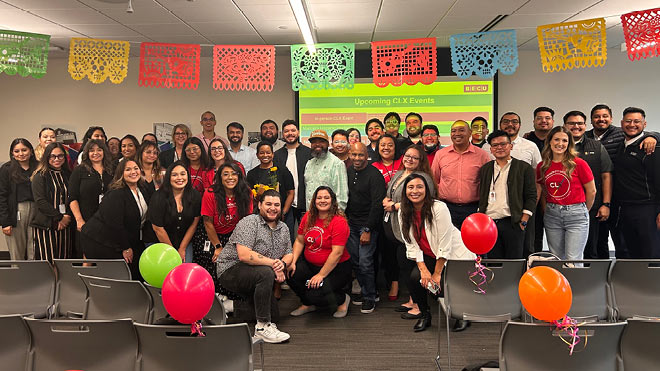 A large group of BECU's Latinx Employee Resource Group (ERG) are gathered together inside at an employee event. The group is standing in front of a screen posing for a photo. There are balloons and decorations in the background.