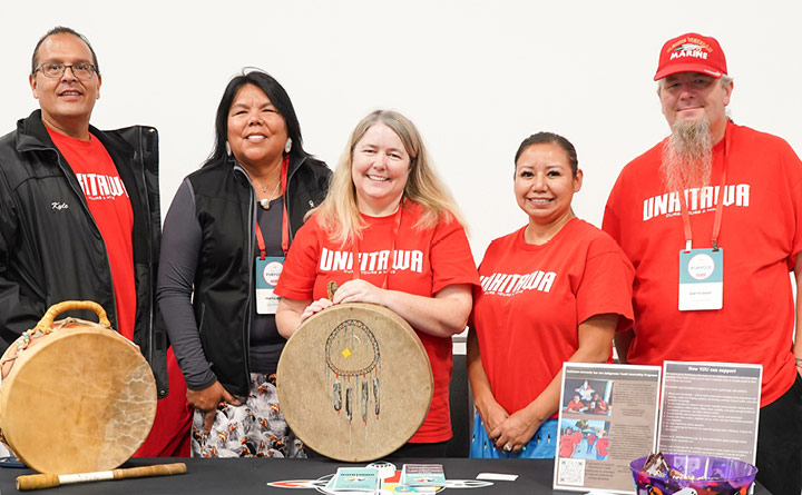 Five members from the Unkitawa nonprofit are standing for a group photo. They are inside at a booth. Four of the individuals are wearing red Unkitawa t-shirts. Several are wearing BECU branded lanyards across their necks. Displayed on the booth are a couple artifacts, informational flyers and papers.
