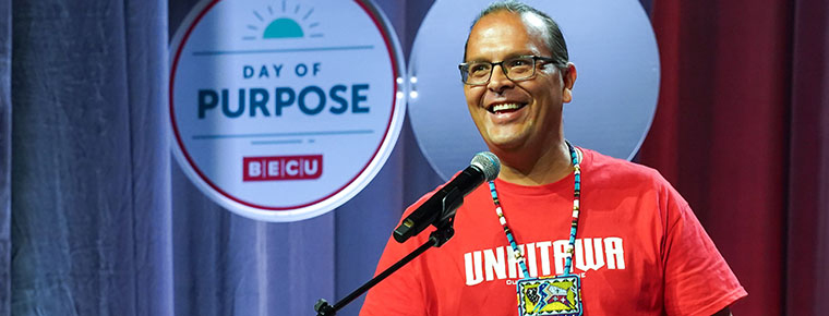 Kyle Schierbeck, project executive for Unkitawa, is standing on a stage in front of microphone smiling. He is wearing a red shirt that says Unkitawa and is wearing a large beaded colorful necklace. Behind him is a BECU sign that says "Day of Purpose BECU."
