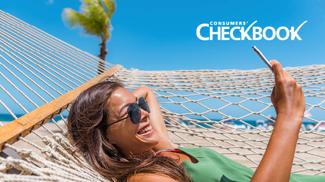 A smiling woman wearing sunglasses and a green top lies in a hammock while looking at her phone. A sandy beach, blue ocean and bright blue sky are in the background. The Consumers' Checkbook logo is in the upper right corner.