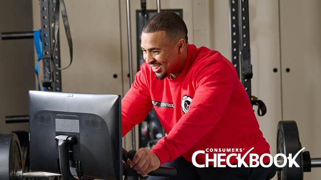 A man is smiling and riding a stationary bike indoors. In the background is fitness equipment. He is looking at a screen while riding the bike. Both of his hands are on the handlebars and he is wearing a red crewneck sweater. The photo has a Consumers' Checkbook logo and the bottom right-hand corner.