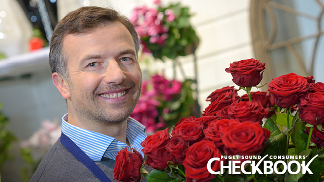 This is an image of a man in a florist shop. He is holding a boutique of red roses. He is smiling and wearing a blue striped collared shirt. In the background are more flower arrangements. In the lower right-hand corner of the photo is the Consumers' Checkbook logo. 