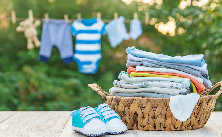 Light blue baby shoes positioned on a wooden deck outside, sitting next to a basket full of folded baby clothes, with baby clothes hanging on a clothesline to dry in background