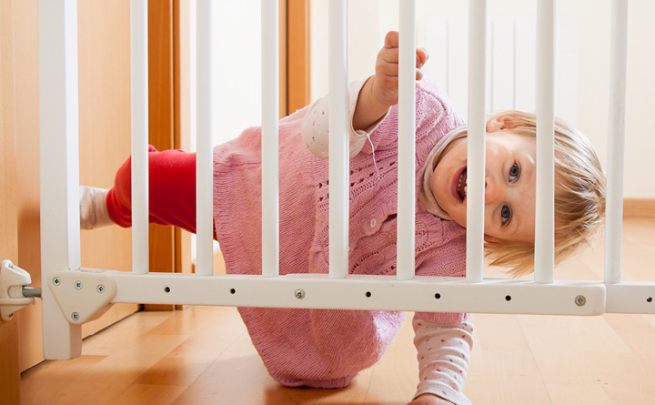 Newborn baby wearing a pink outfit, smiling while looking through a baby gate set up inside a home.