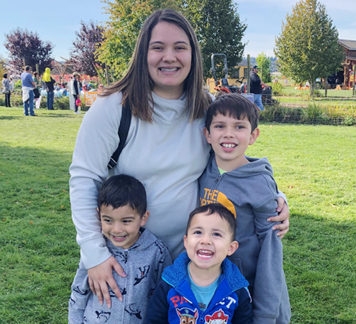 Marybeth and her children smile for a picture on a sunny day at a park. Green grass, trees and blue sky are in the background.