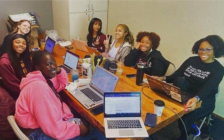 A group of seven women are sitting around a table, smiling at the camera. The table has laptops and coffee on it.